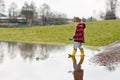 Happy little kid boy in yellow rain boots playing with paper ship boat by huge puddle on spring or autumn day. Active Royalty Free Stock Photo