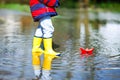 Happy little kid boy in yellow rain boots playing with paper ship boat by huge puddle on spring or autumn day Royalty Free Stock Photo