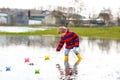 Happy little kid boy in yellow rain boots playing with paper ship boat by huge puddle on spring or autumn day Royalty Free Stock Photo