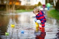 Happy little kid boy in yellow rain boots playing with paper ship boat by huge puddle on spring or autumn day Royalty Free Stock Photo