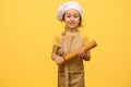 Happy little preschool girl with rolling pin, dressed as chef pastry, smiling looking at camera, yellow background Royalty Free Stock Photo