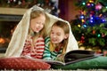 Happy little sisters reading a story book together by a fireplace in a cozy dark living room on Christmas eve. Royalty Free Stock Photo