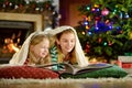 Happy little sisters reading a story book together by a fireplace in a cozy dark living room on Christmas eve.