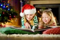 Happy little sisters reading a story book together by a fireplace in a cozy dark living room on Christmas eve. Royalty Free Stock Photo