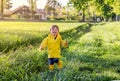Happy little smiling boy in bright yellow raincoat and rubber boots running through the field with green grass Royalty Free Stock Photo