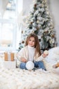 Happy little smiling girl with gift on Christmas Eve sit on bed. Child opens New year gift near christmas tree with glowing lights Royalty Free Stock Photo