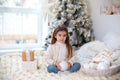 Happy little smiling girl with gift on Christmas Eve sit on bed. Child opens New year gift near christmas tree with glowing lights Royalty Free Stock Photo