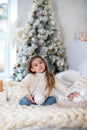 Happy little smiling girl with gift on Christmas Eve sit on bed. Child opens New year gift near christmas tree with glowing lights Royalty Free Stock Photo