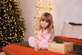 Happy little smiling girl in pajamas with gift on xmas Eve lies on bed. child opens New year gift at home near christmas tree with Royalty Free Stock Photo