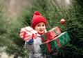 Happy little smiling girl in red cap with Christmas gift boxes standing near by spruce. Holiday celebrate concept