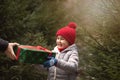 Happy little smiling girl in red cap takes a given box with a gift for Christmas. Kid standing near by spruce. Holiday celebrate