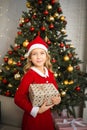 Happy little smiling girl in santa hat with christmas gift box stanging near Christmas tree. Royalty Free Stock Photo