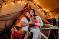 Happy lovely couple relaxing in glamping on summer evening and drinking wine near cozy bonfire. Luxury camping tent for outdoor