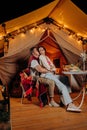 Happy lovely couple relaxing in glamping on summer evening and drinking wine near cozy bonfire. Luxury camping tent for outdoor