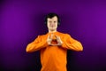 Happy male in headphones shows the gesture of the heart. A smiling guy in an orange T-shirt on a purple background shows a heart Royalty Free Stock Photo