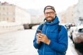 Happy man with beard wearing eyeglasses dressed in blue anorak holding backpack and mobile having happy look travelling in city wa