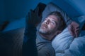 Happy man with blue eyes lying on bed late at night in dark light networking on mobile phone or online dating smiling relaxed Royalty Free Stock Photo