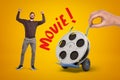 Happy man in casual clothes and hand holding hand truck with film reel and `Movie` sign on yellow background.