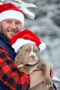 Happy man holding lovely dog in his hands wearing in a Santa hat in snowy forest. Smiling boy hugging adorable puppy in Royalty Free Stock Photo