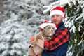 Happy man holding lovely dog in his hands wearing in a Santa hat in snowy forest. Smiling boy hugging adorable puppy in Royalty Free Stock Photo