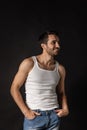 Portrait of young handsome bearded sad man in white undershirt and jeans posing isolated on dark background. Royalty Free Stock Photo