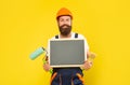 happy man renovator in helmet and work clothes hold paint roller brush and blackboard