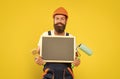 happy man renovator in helmet and work clothes hold paint roller brush and blackboard