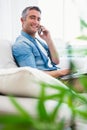 Happy man sitting on couch phoning and using laptop Royalty Free Stock Photo