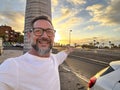Happy man taking portrait selfie picture against a colorful sunset on the street with car and roads. Travel and summer vacation