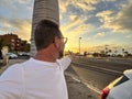 Happy man taking portrait selfie picture against a colorful sunset on the street with car and roads. Travel and summer vacation