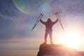 A happy man on top of a mountain with crutches, raising his hands up, opposite a dramatic sky at dawn.