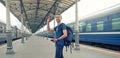 Happy man tourist with backpack stand on railway station platform, greeting friends or saying goodbye, waving his hand. Travel by