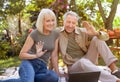 Happy married senior couple using laptop computer and video calling family, sitting on picnic blanket in garden Royalty Free Stock Photo