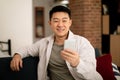 Happy mature asian man in casual watching video or chatting on smartphone, resting on sofa in living room interior