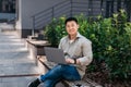 Happy mature asian man working on laptop computer outdoors, sitting on bench in urban area and using pc Royalty Free Stock Photo