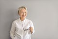 Happy mature business woman standing by grey copy space wall, holding glasses and smiling Royalty Free Stock Photo