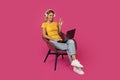 Happy mature caucasian blonde woman in casual and headphones sits on armchair, waving hand at laptop Royalty Free Stock Photo
