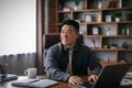 Happy mature chinese man at table working with laptop in home office interior and looking at empty space Royalty Free Stock Photo