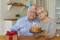 Happy mature couple in cone hats blowing out candles on cake celebrating birthday at home.