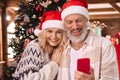 Happy mature couple in santa hats using cell phone near Christmas tree at home. Royalty Free Stock Photo
