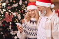 Happy mature couple in santa hats using cell phone near Christmas tree at home. Royalty Free Stock Photo