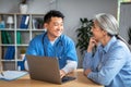 Happy mature japanese male doctor shows laptop, talk on consultation to senior caucasian woman patient in clinic