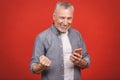 Happy mature senior man in casual using smartphone and making winner gesture isolated over red background. Using phone