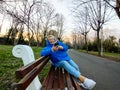 Happy mature woman in blue hoodie looking at mobile, sitting on bench in park at sunset. Royalty Free Stock Photo