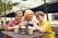 Happy mature women sitting at table with coffee and laughing while watching video on smartphone
