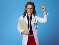 Happy medical doctor woman with clipboard rising finger on blue
