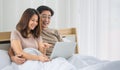Happy middle age Asian couple dressed casually sitting relaxed on a bed at home enjoy looking at a laptop computer. Warmth,