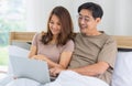 Happy middle age Asian couple dressed casually sitting relaxed on a bed at home enjoy looking at a laptop computer. Warmth,