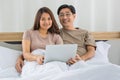 Happy middle age Asian couple dressed casually sitting relaxed on a bed at home enjoy looking at a laptop computer. Warmth,