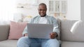 Happy middle-aged black man using laptop while resting on sofa in living room, panorama Royalty Free Stock Photo